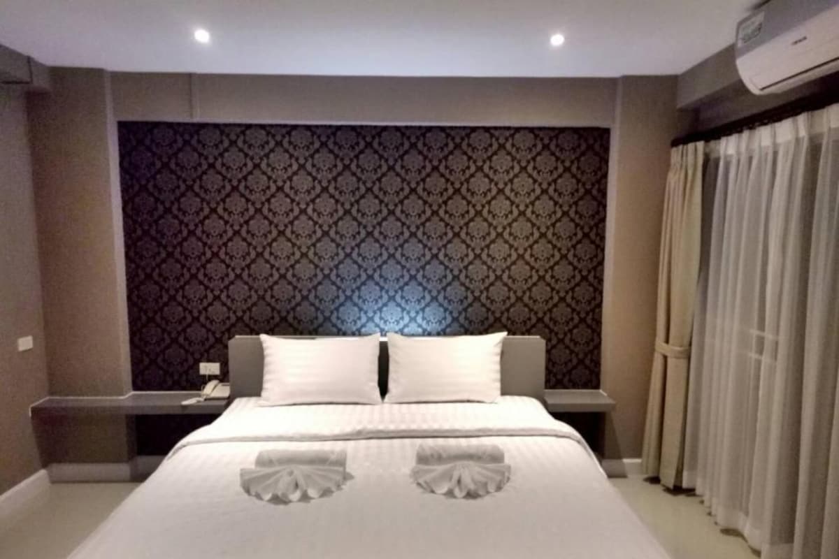 A hotel room with a large bed, a patterned headboard, a bedside table with a lamp, and a window with curtains.