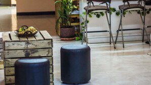 A reception area with a counter, two bar stools, a potted plant, a wooden chest, and a person seated behind the counter. - Karma Home Hostel (Phitsanulok)