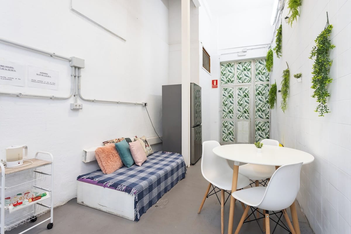 Una habitación pequeña y funcional con cama, una mesa blanca con sillas, una pizarra con texto y plantas verdes en la pared.