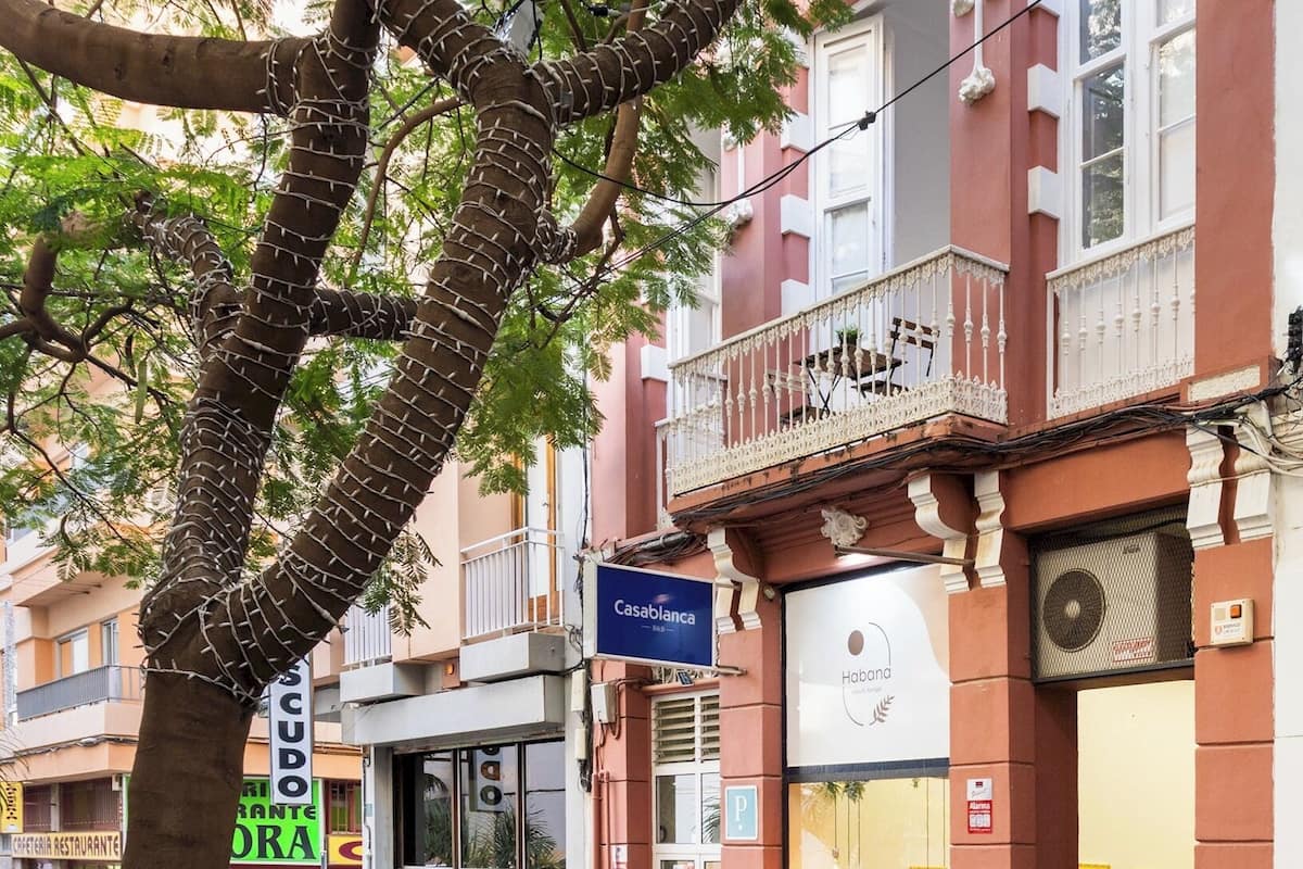 Vista callejera con un árbol, un edificio con balcón y una tienda con letrero.
