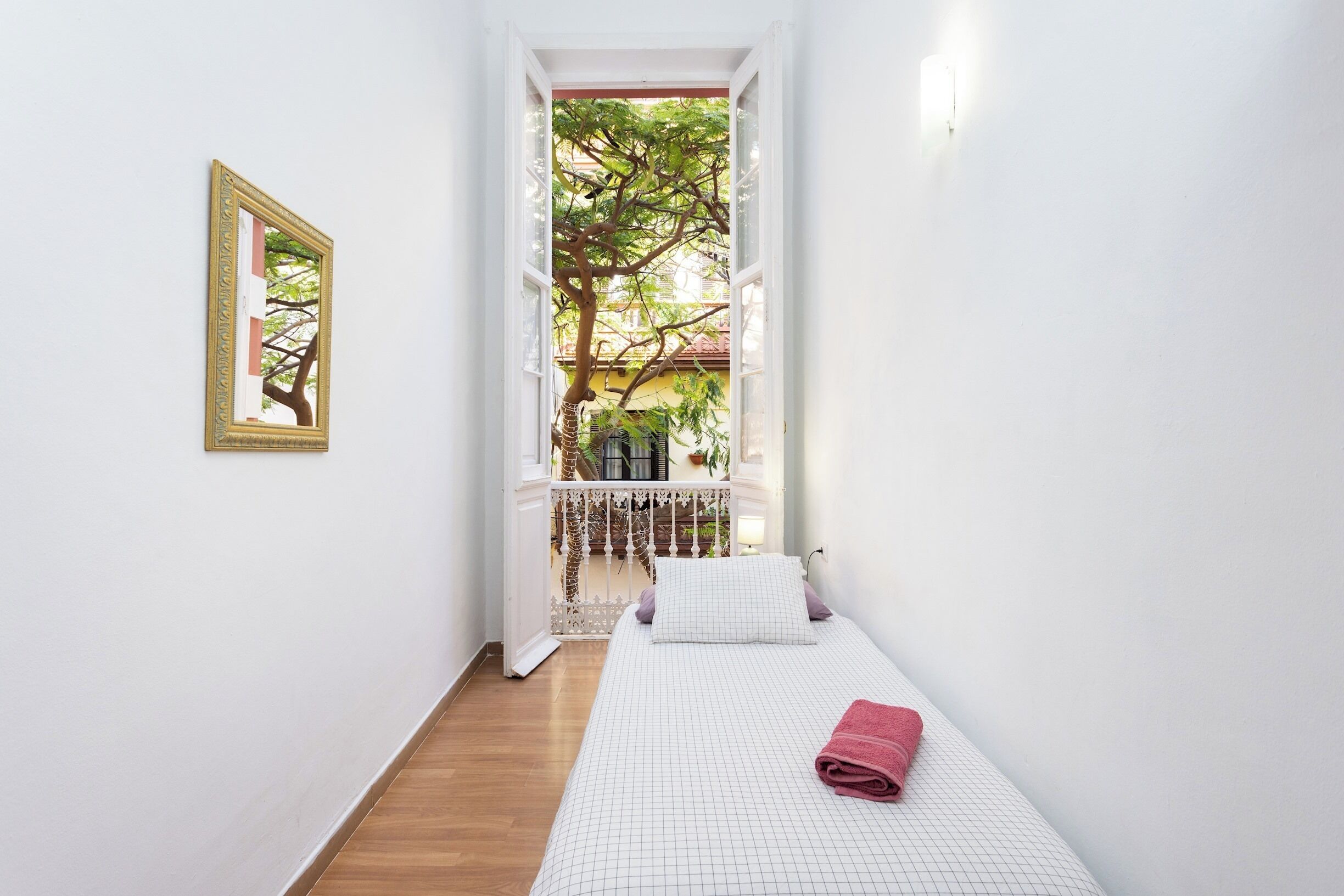 A narrow hallway leading to a bed with a white grid-patterned bedspread and a pink towel, a framed picture on the wall, and a view of a balcony with a tree.