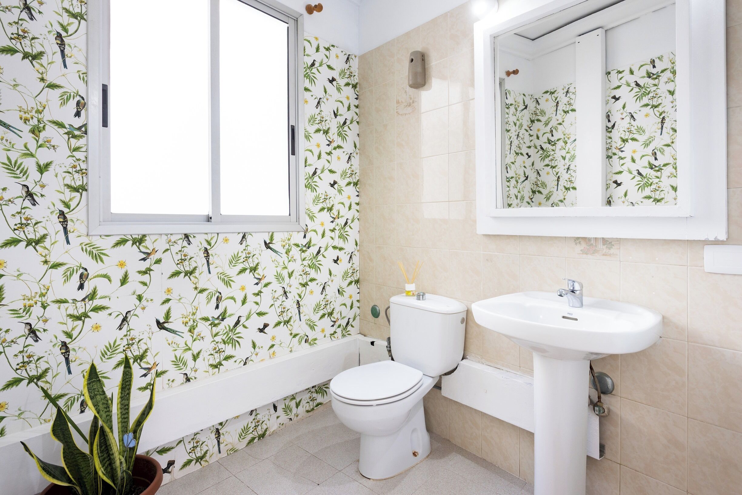 Un baño con inodoro, lavabo y bañera, con papel tapiz floral y una ventana.