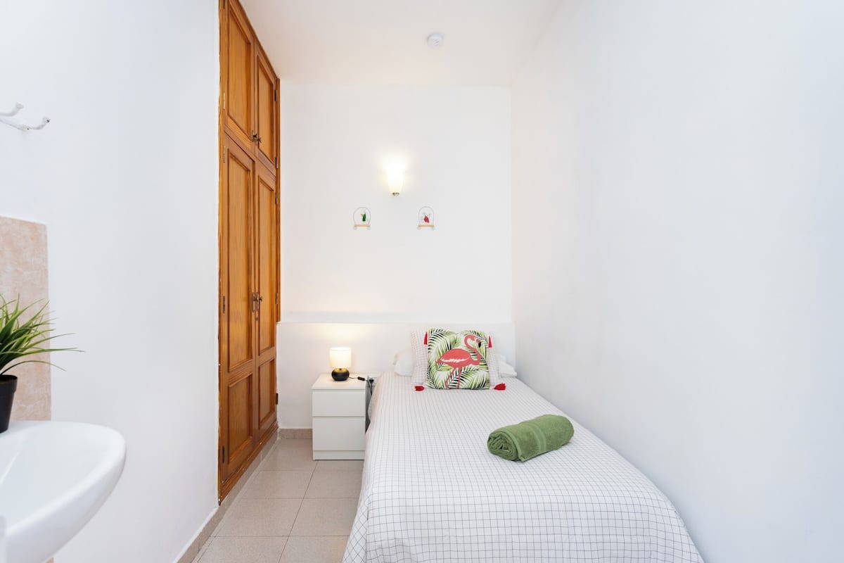 Una habitación pequeña y limpia con una cama, una mesita de noche, un armario de madera y una pared blanca.