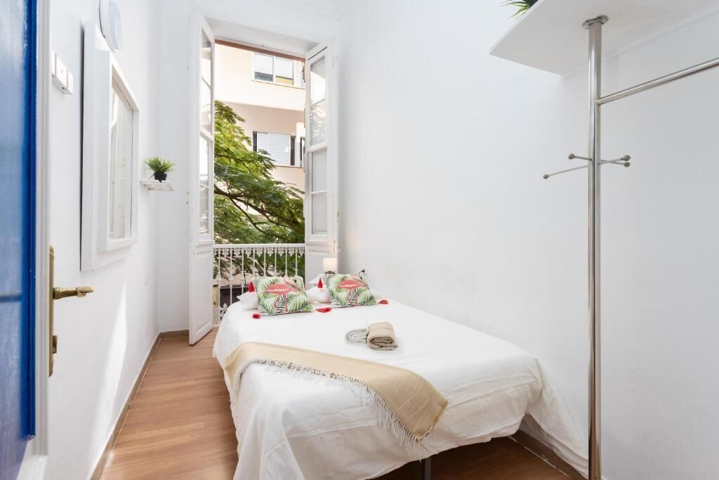 Una habitación estrecha con cama, una puerta blanca y vistas a un balcón con plantas.