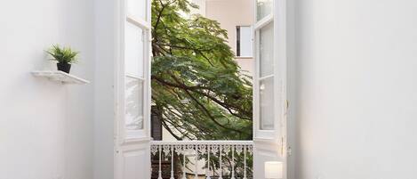 Habitación de hotel con cama, ventana, una pequeña estantería con una planta y un balcón con barandilla.