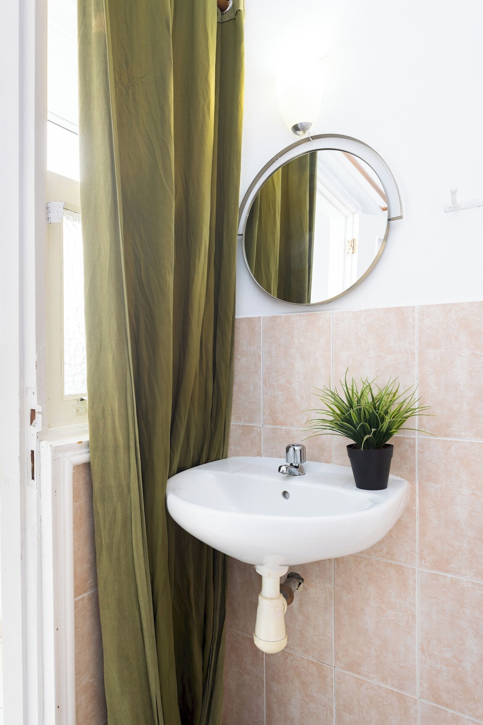 Un bagno con un lavandino bianco, uno specchio rotondo, una pianta in vaso e una tenda verde.