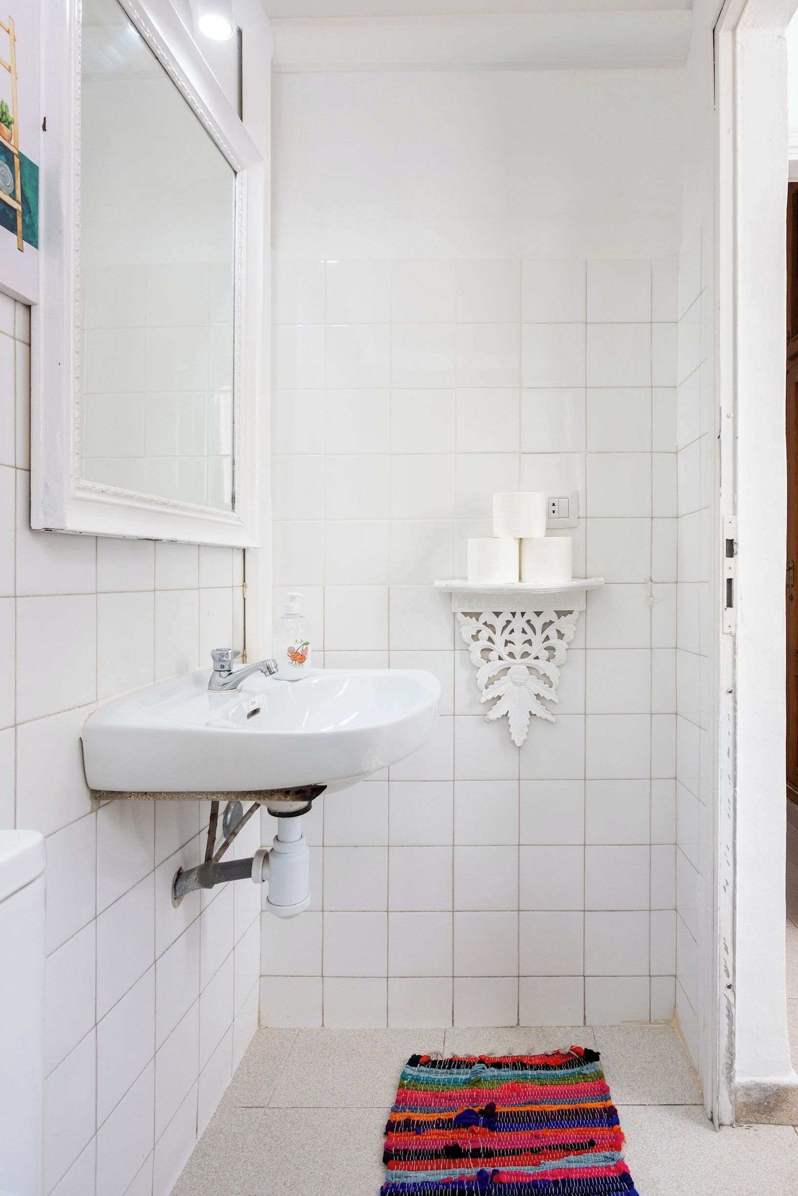 A bathroom with a white sink, mirror, and toilet. There is a shelf with toiletries and a colorful rug on the floor.