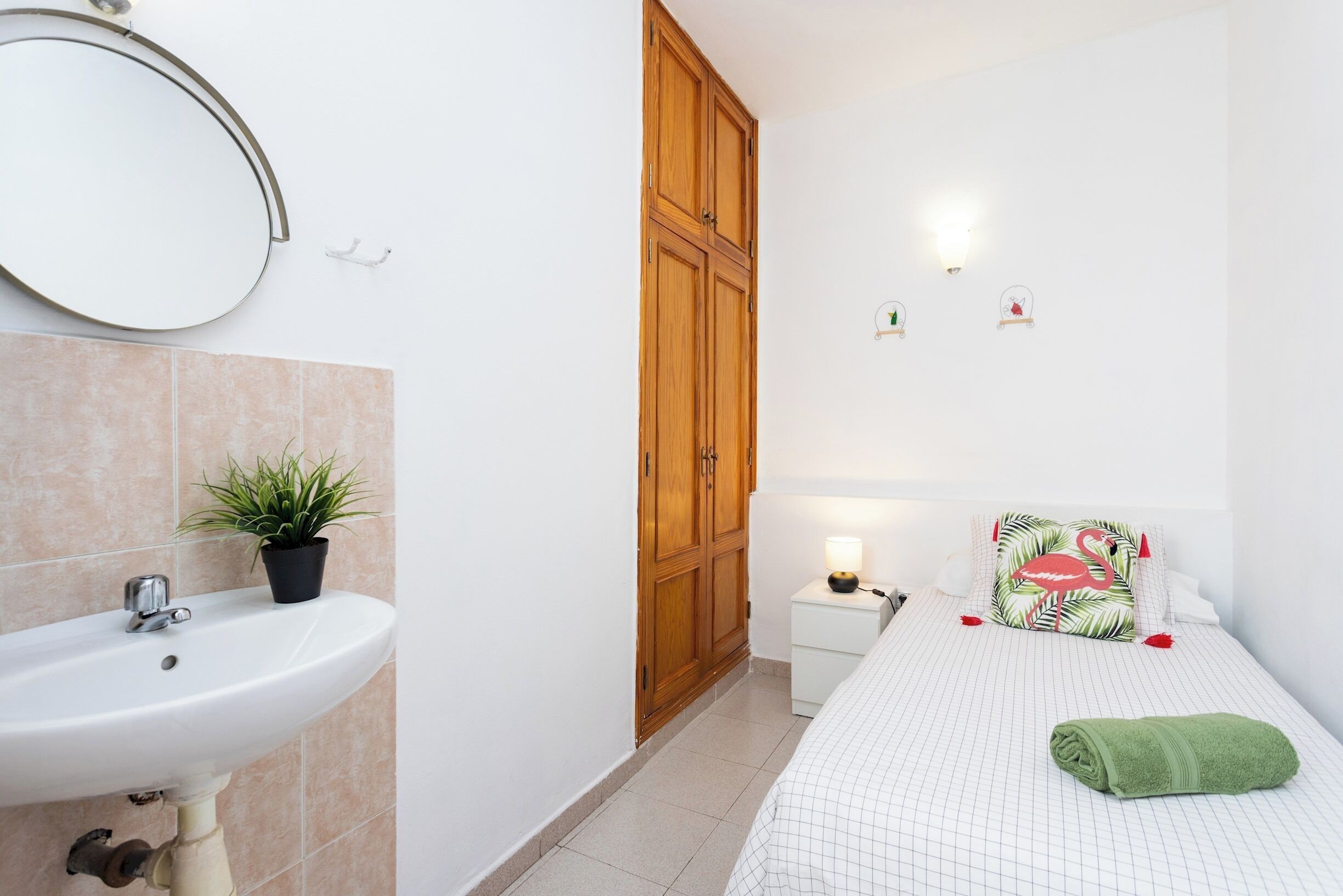 Een slaapkamer met een bed, nachtkastje, lamp, spiegel, houten kledingkast en een kamerplant.