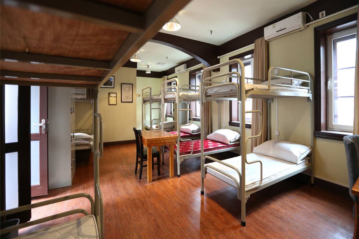 Une chambre de dortoir avec des lits superposés, une table en bois, des chaises et une fenêtre avec des rideaux.