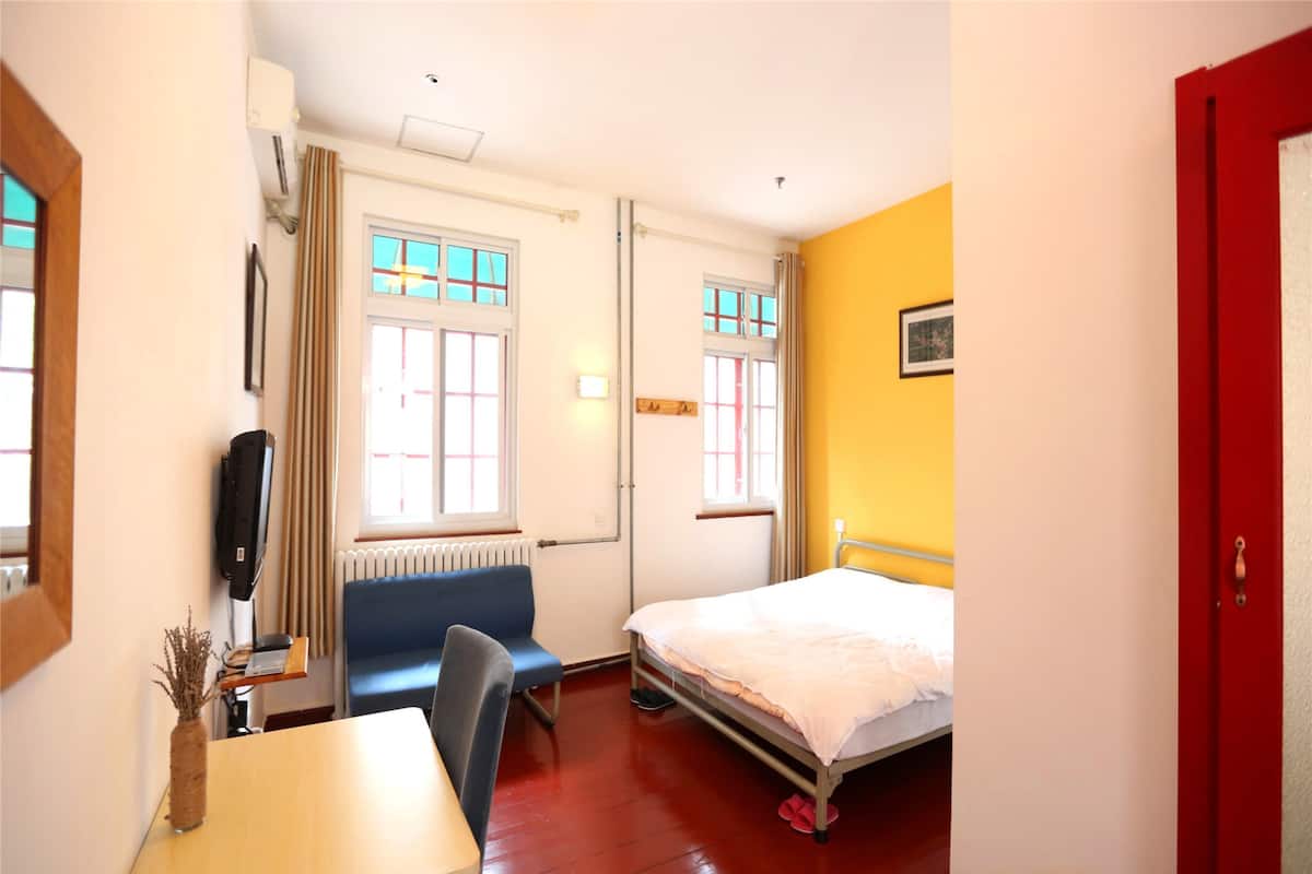 Une chambre d’hôtel avec un lit, un bureau, une chaise, un radiateur et deux fenêtres.