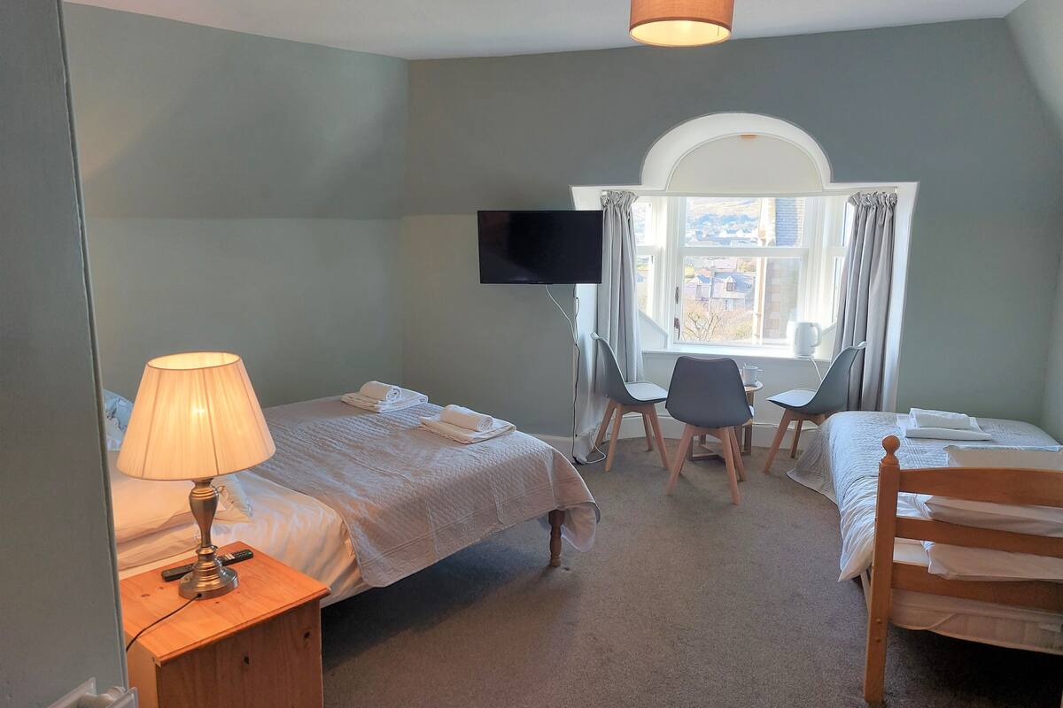 Ein Hotelzimmer mit zwei Betten, einem Schreibtisch, Stühlen, einem Fernseher und einem Fenster mit Vorhängen.