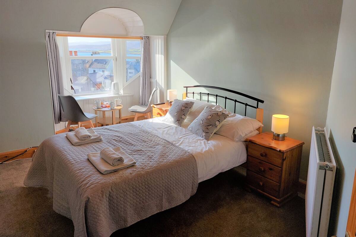 Ein Schlafzimmer mit Bett, Nachttisch, Lampe, Stuhl und einem Fenster mit Vorhängen.