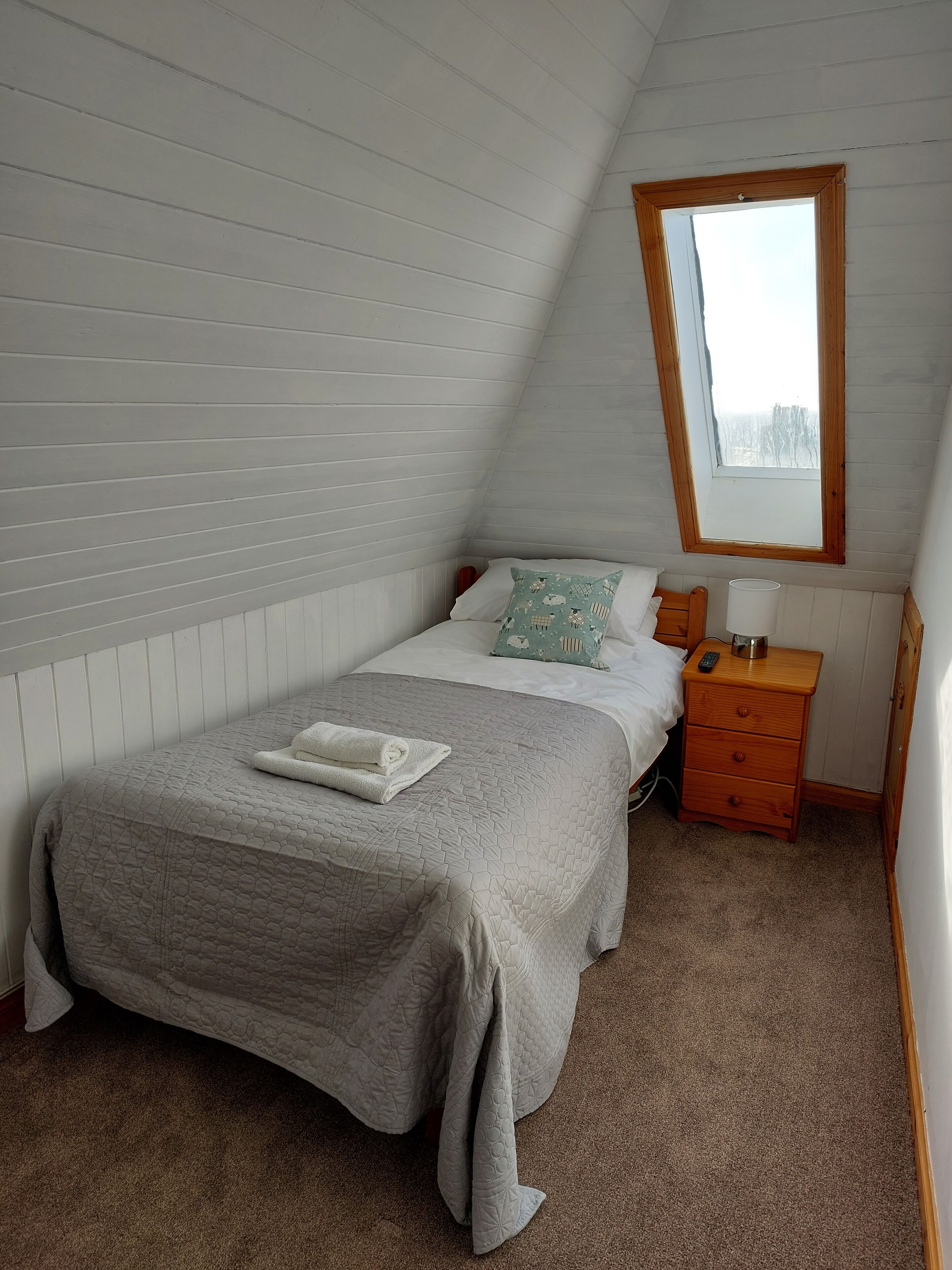 A single bed with a quilt, a bedside table with a lamp, and a window in a room with sloped ceilings.