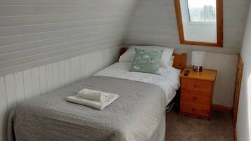 A single bed with a quilt, a bedside table with a lamp, and a window in a room with sloped ceilings.