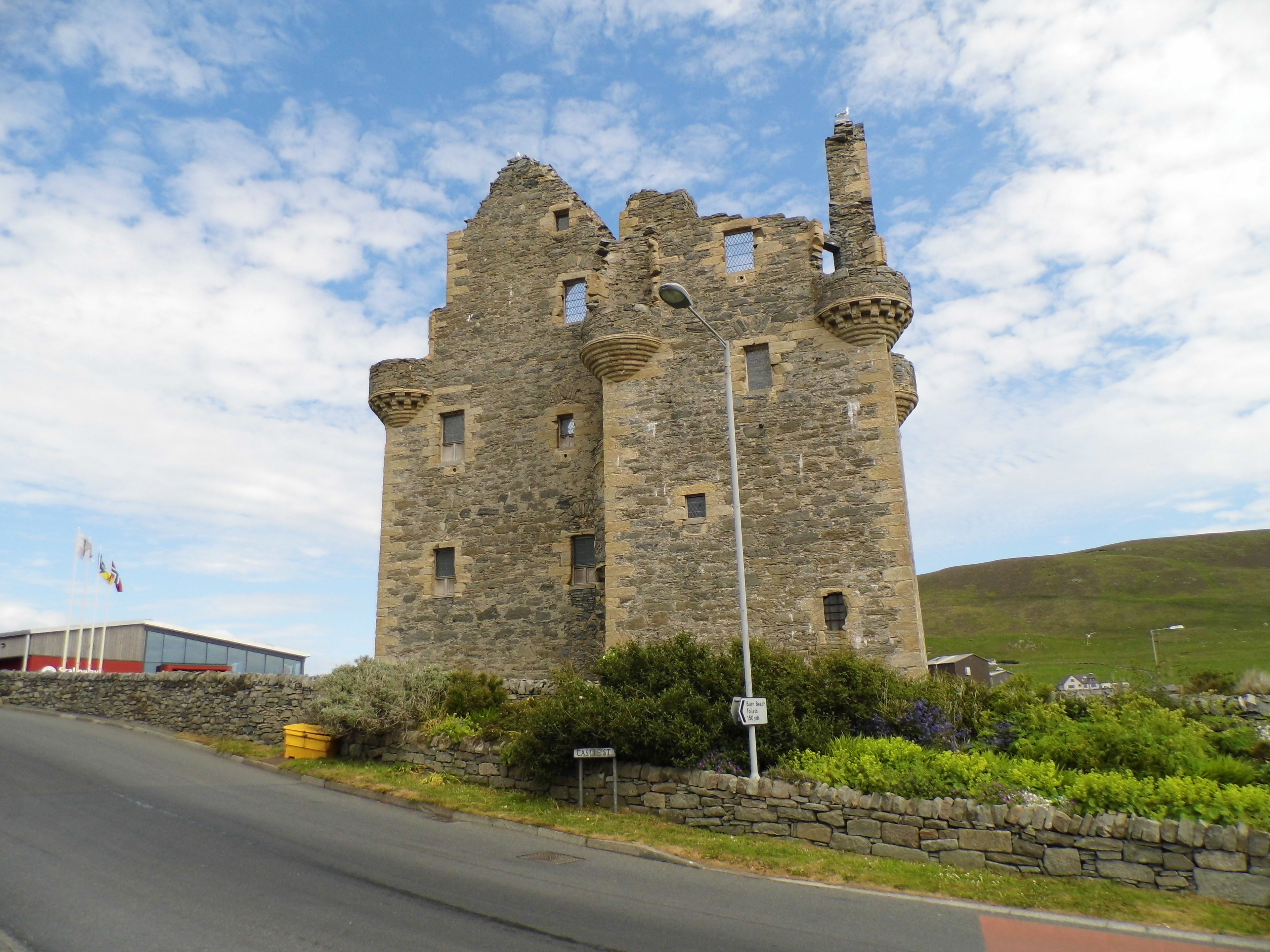A historic stone castle with multiple towers and battlements, situated beside a road with a stone wall and greenery.