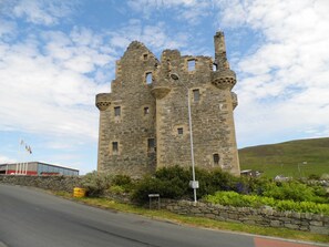 A historic stone castle with multiple towers and battlements, situated beside a road with a stone wall and greenery. - Eddlewood Guest House (Lerwick)