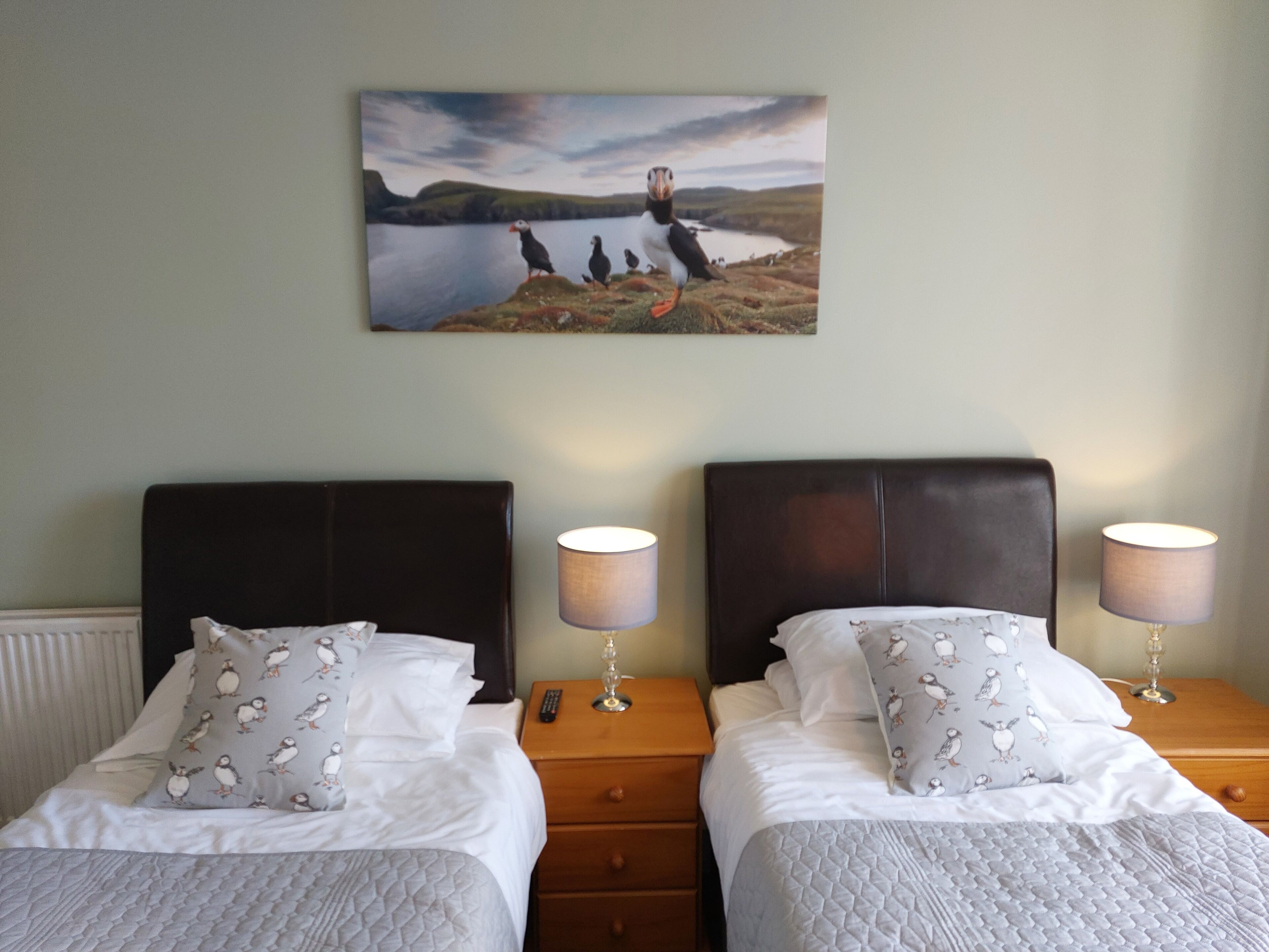 Habitación de hotel con dos camas, cada una con un cojín estampado con pájaros, una mesita de noche con lámpara y un cuadro de aves en la pared.