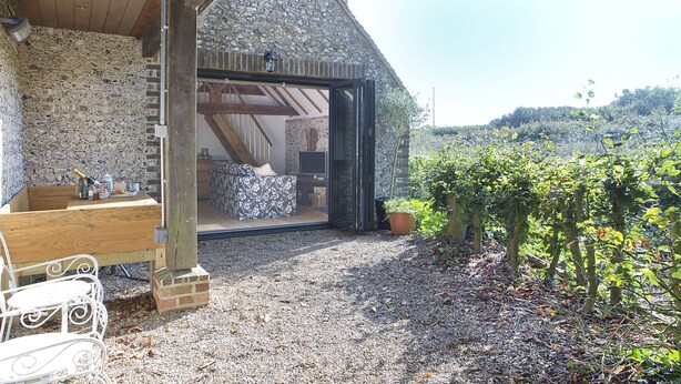 A rustic stone building with a wooden bench and table outside, a gravel path leading to a glass door, and a view of a vineyard in the background. - Shepherds Barn (Chichester)