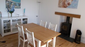 A dining area with a wooden table and white chairs, a white cabinet with glass doors, a painting of a cow, and a wood-burning stove. - Bluebell Bank (Dumfries)