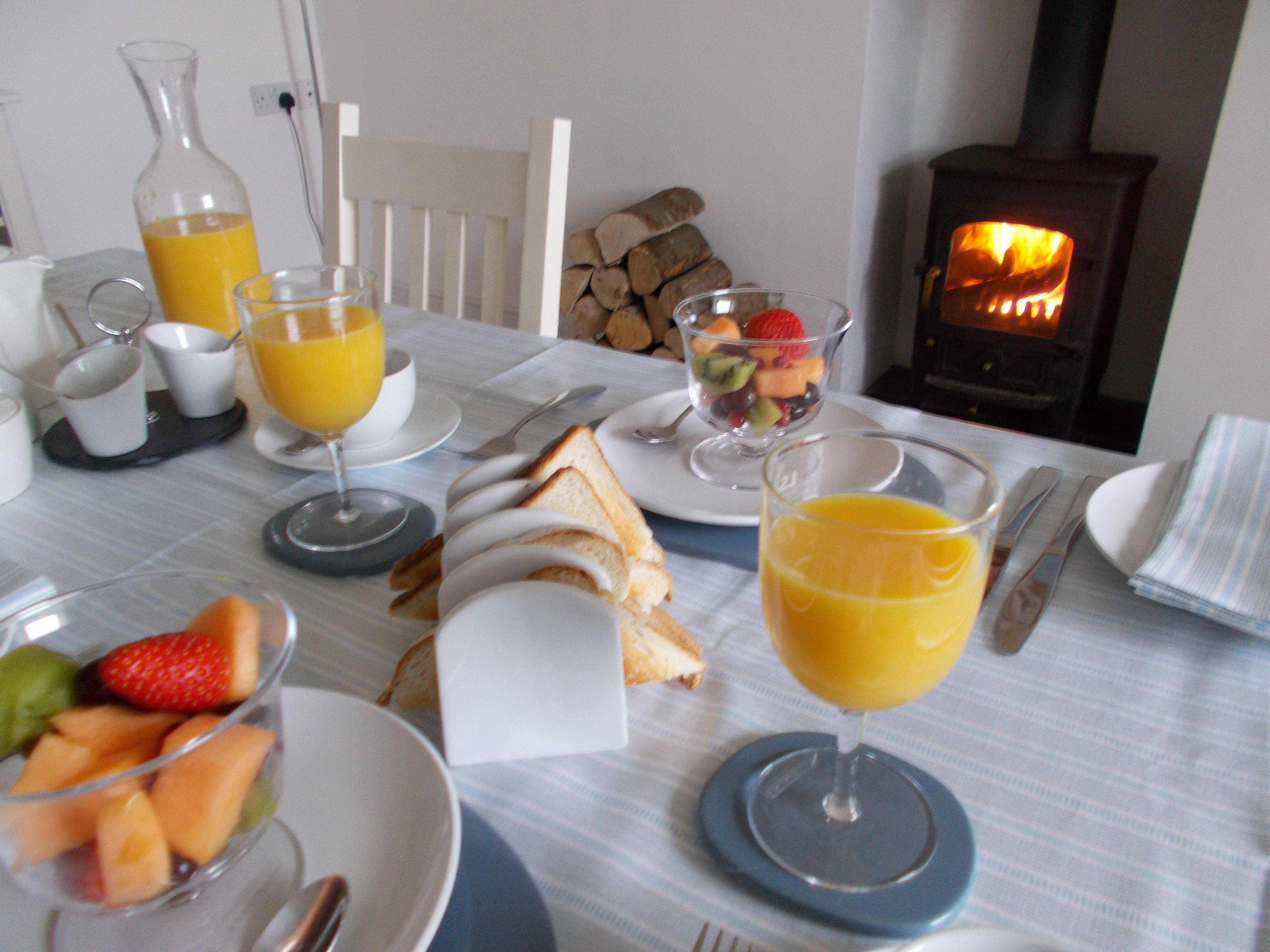 A breakfast setting with toast, fruit, and orange juice on a table, with a wood-burning stove in the background.
