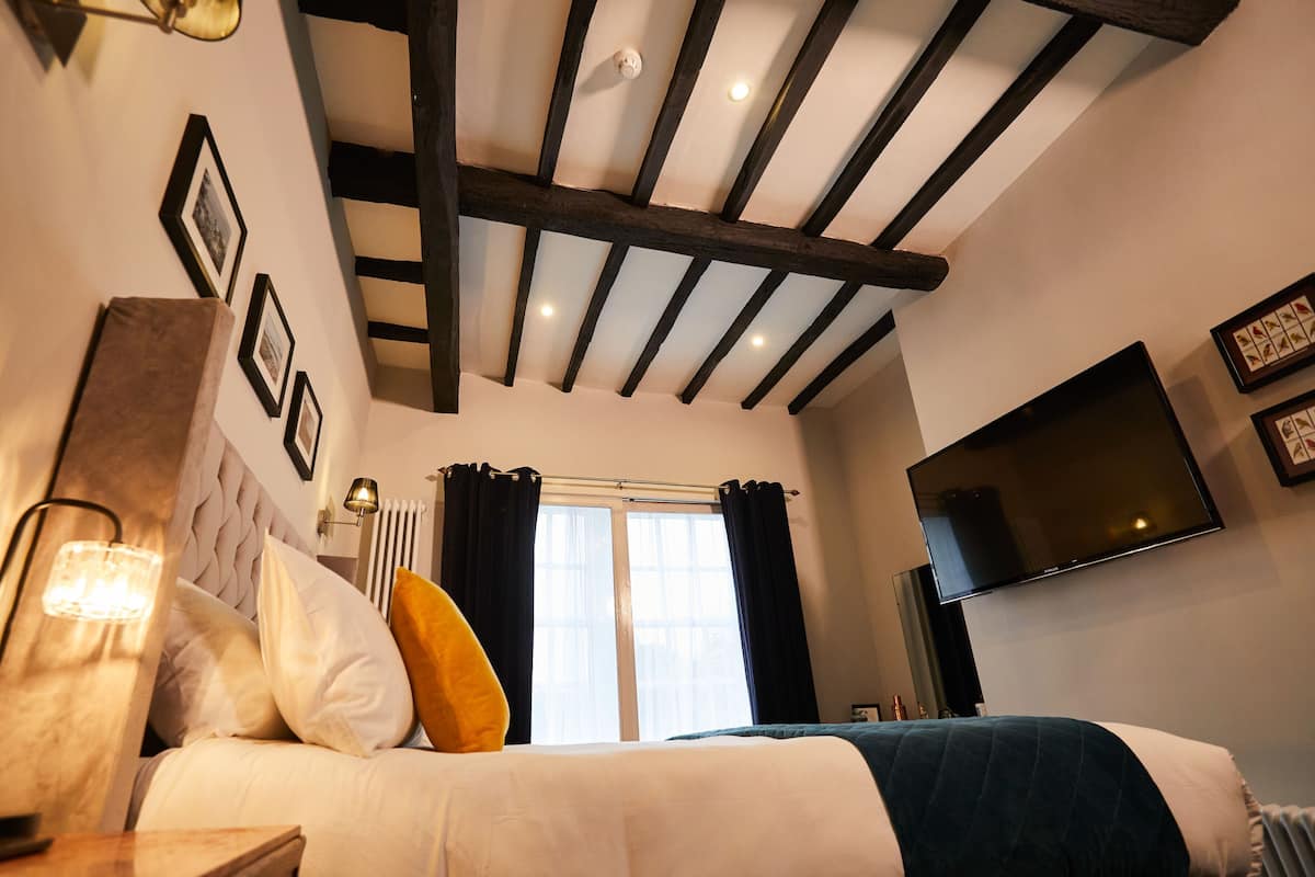 A bedroom with a bed, a TV, a window with curtains, and a ceiling with exposed beams.