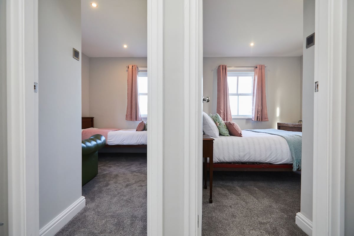 A bedroom with two single beds, a window with curtains, a carpeted floor, and a door leading to another room.