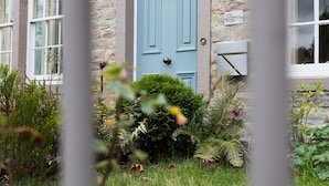 Un edificio de piedra con una puerta azul, un cartel que indica que es un hotel, y un jardín bien cuidado.