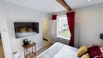 Un dormitorio con una cama, un televisor montado en la pared, una mesita de noche con cafetera y una ventana con cortinas rojas, que ofrece vistas al exterior.
