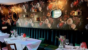 Interior de un restaurante con papel tapiz floral, asientos tapizados en verde oscuro y mesas dispuestas para comer.