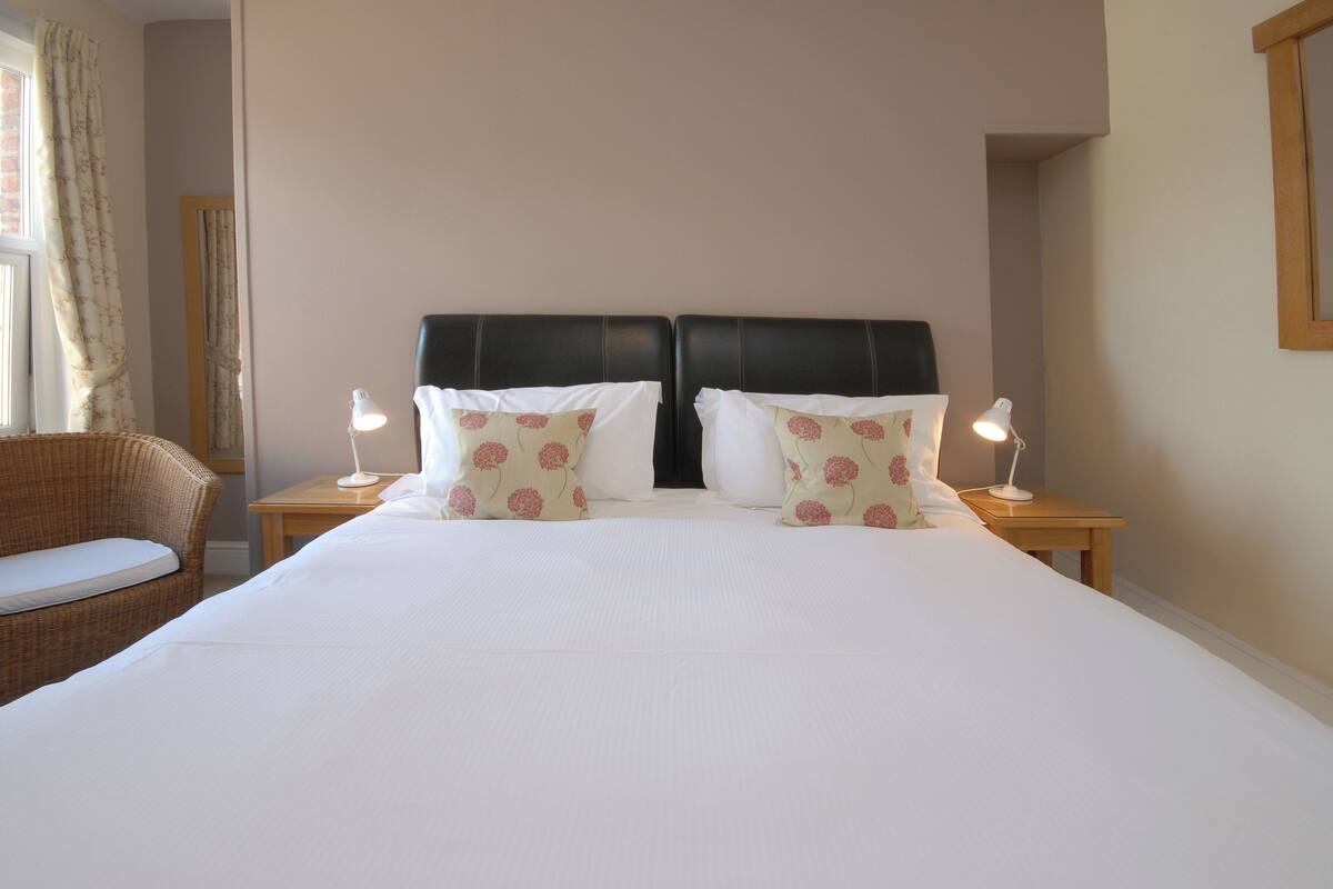 A neatly made bed with white linens and floral pillows, flanked by two bedside tables with lamps, in a room with a wicker chair and a plain wall.
