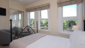 A bedroom with a bed, two windows with curtains, a chair, and a bedside table with a lamp. - Abbey Guest House (York)