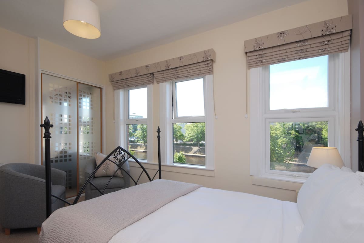A bedroom with a bed, two windows with curtains, a chair, and a bedside table with a lamp.