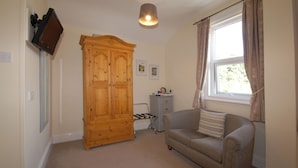 A room with a wooden wardrobe, a grey sofa with a cushion, a small desk with a chair, and a window with curtains. - Abbey Guest House (York)