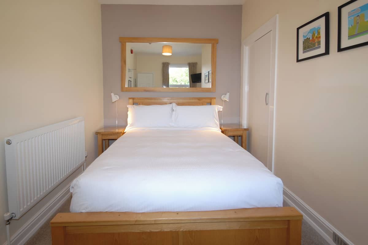 A neatly made bed in a room with a wooden headboard, a bedside table with a lamp, a radiator, and a large mirror above the bed.