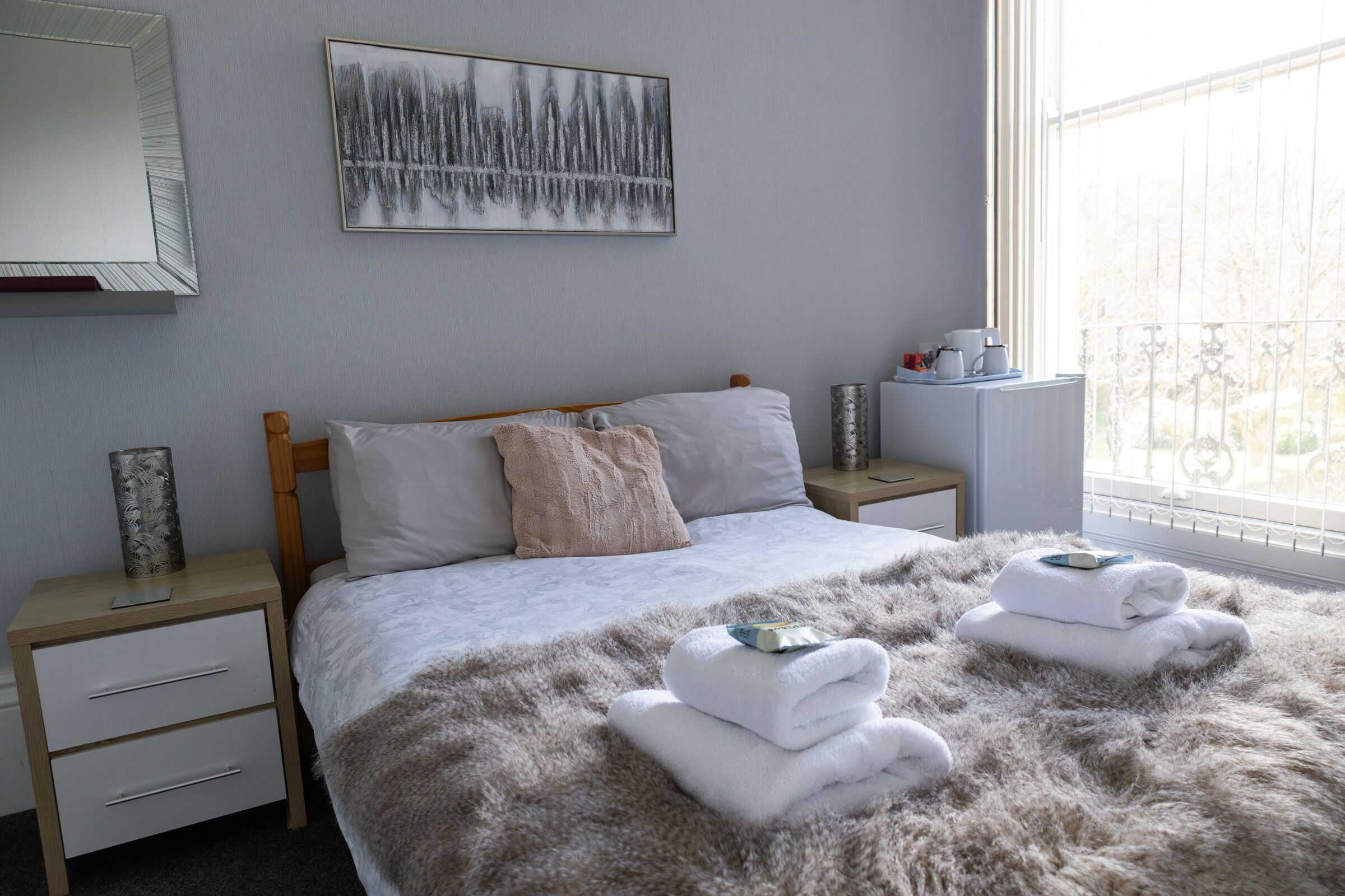 A bedroom with a bed, a nightstand, a bedside table, a window with a view, and a painting on the wall.