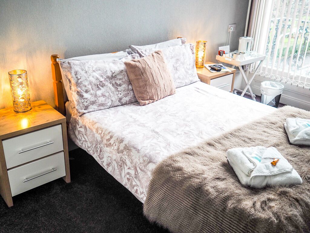 Ein ordentlich bezogenes Bett mit einem gemusterten Überwurf, zwei Kissen und einer flauschigen Decke. Zwei Nachttischlampen, ein kleiner Tisch mit Lampe und ein Fenster mit Vorhängen sind vorhanden.