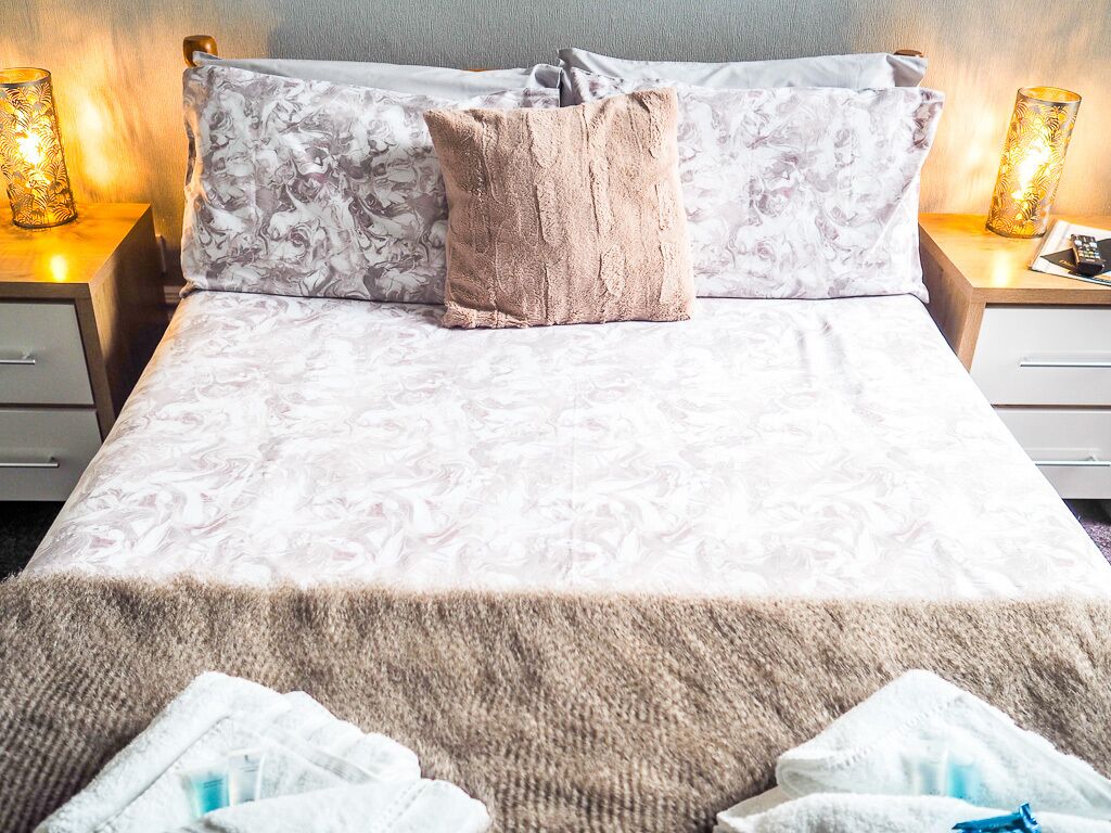 A neatly made bed with a patterned bedspread, a fluffy beige throw blanket, and two pillows. There are two bedside lamps with decorative shades on either side of the bed.