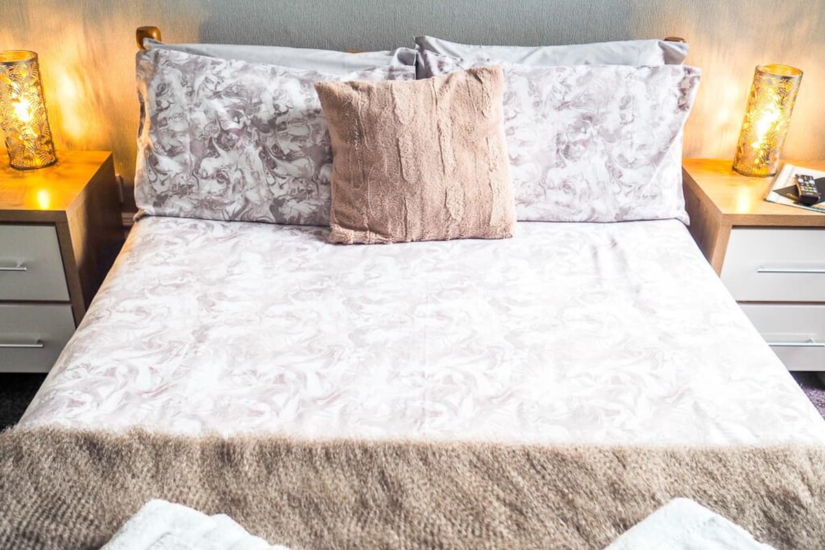 A neatly made bed with a patterned bedspread, a fluffy beige throw blanket, and two pillows. There are two bedside lamps with decorative shades on either side of the bed.