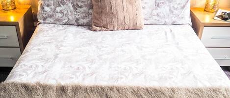 A neatly made bed with a patterned bedspread, a fluffy beige throw blanket, and two pillows. There are two bedside lamps with decorative shades on either side of the bed.