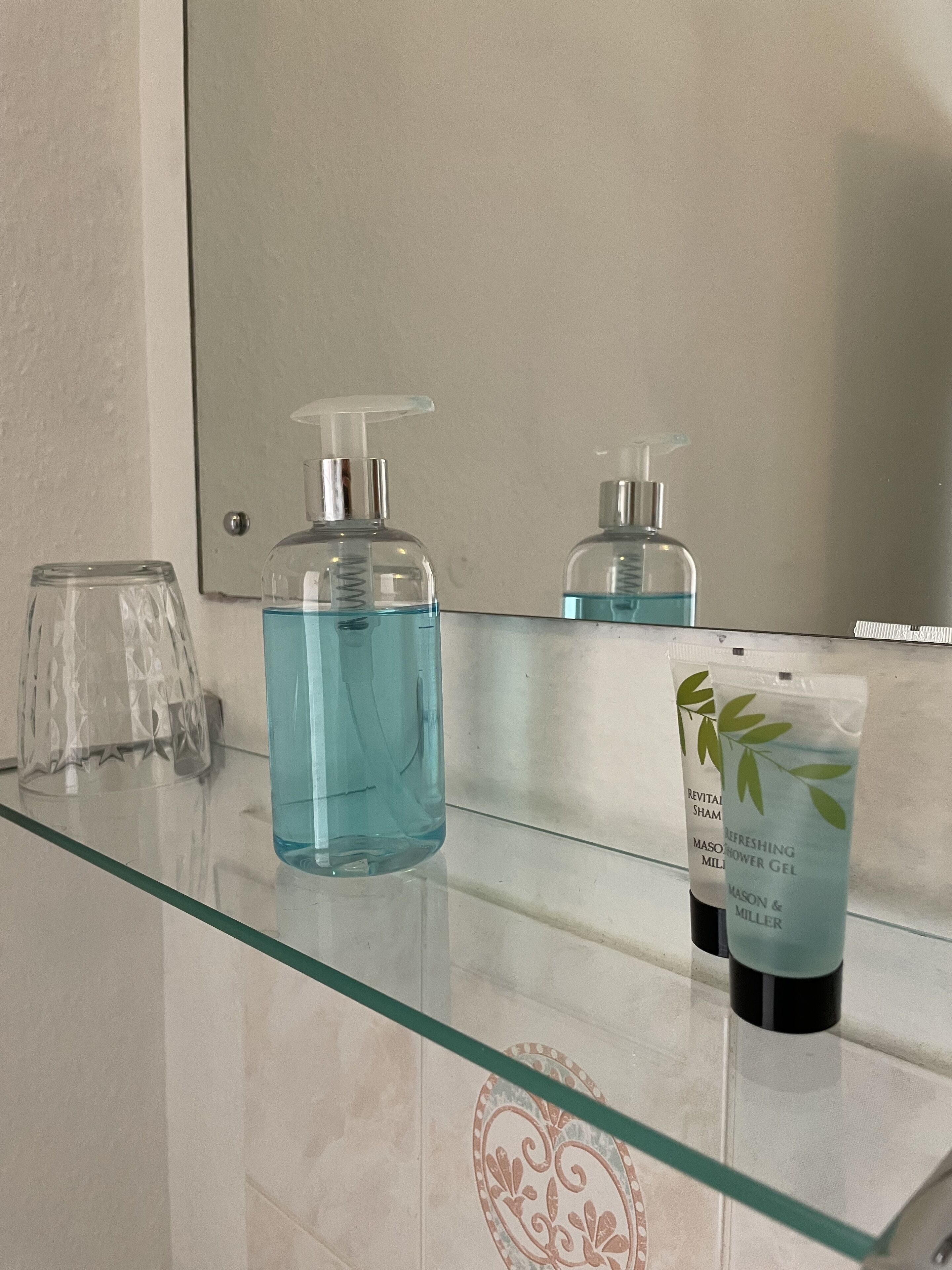 A bathroom with a glass countertop, a large bottle of blue liquid soap, a smaller bottle of shower gel, and a glass.