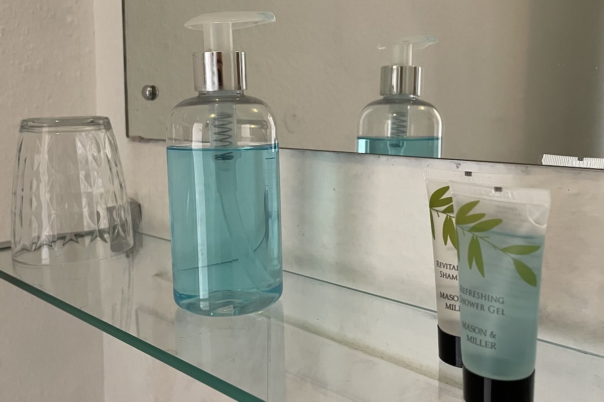 A bathroom with a glass countertop, a large bottle of blue liquid soap, a smaller bottle of shower gel, and a glass.