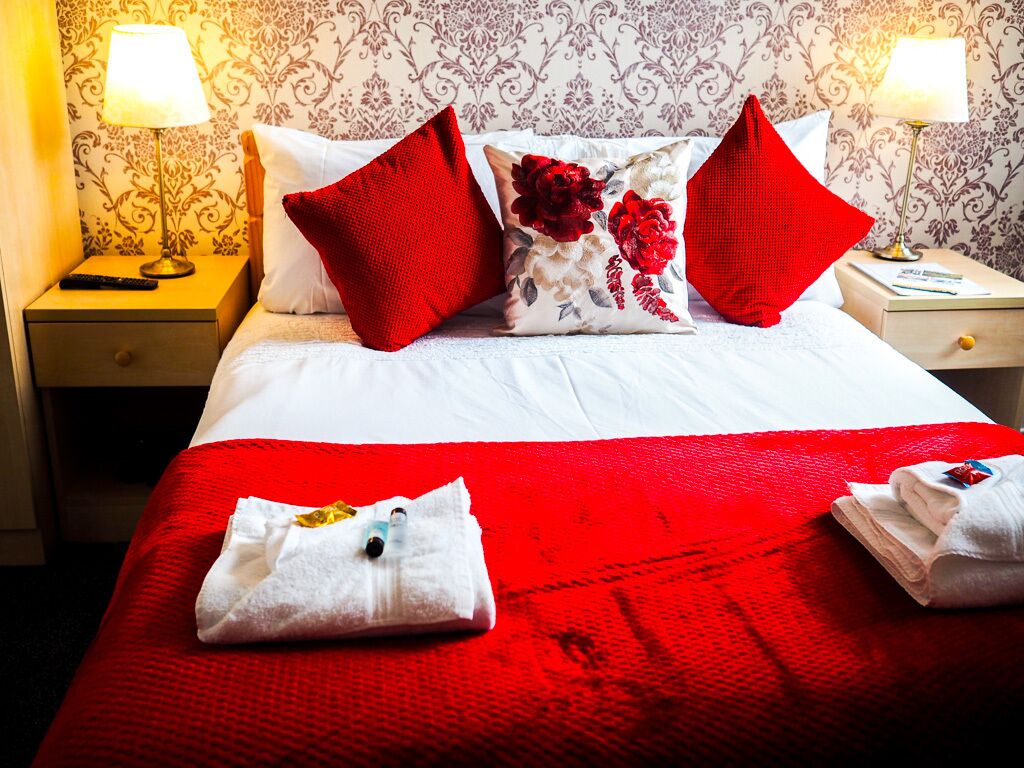 A hotel room with a neatly made bed, red and white bedding, and a floral pillow.