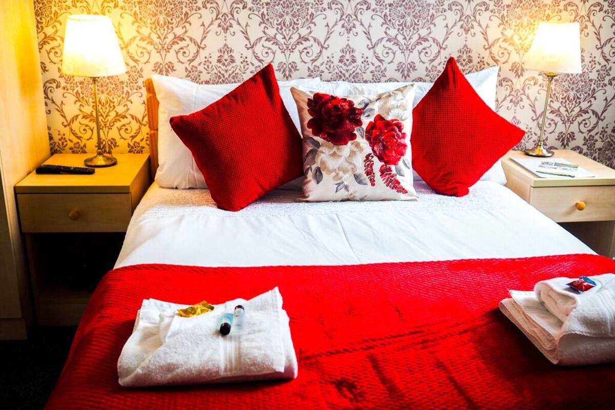 A hotel room with a neatly made bed, red and white bedding, and a floral pillow.