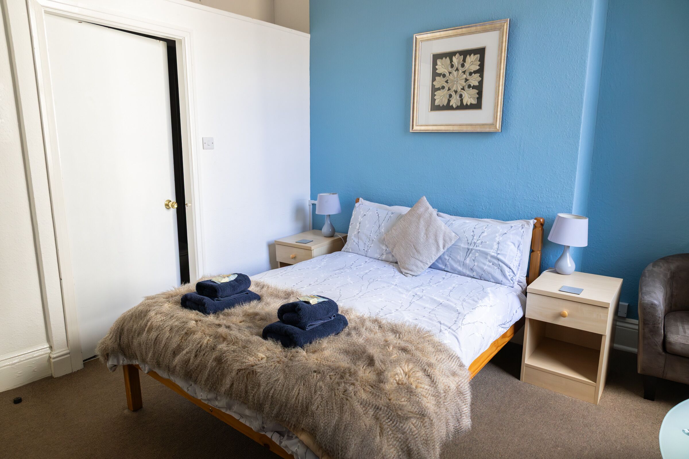Ein Schlafzimmer mit einem Bett, zwei Nachttischen, einem Schreibtisch, einem Sessel, einer Tür und einem gerahmten Bild an der Wand.