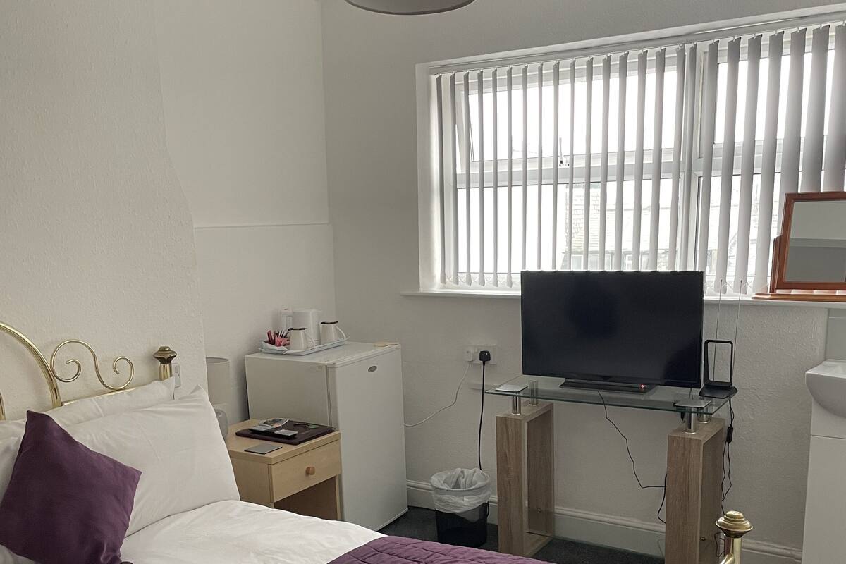 A hotel room with a bed, a desk with a television, a window with blinds, and a trash bin.