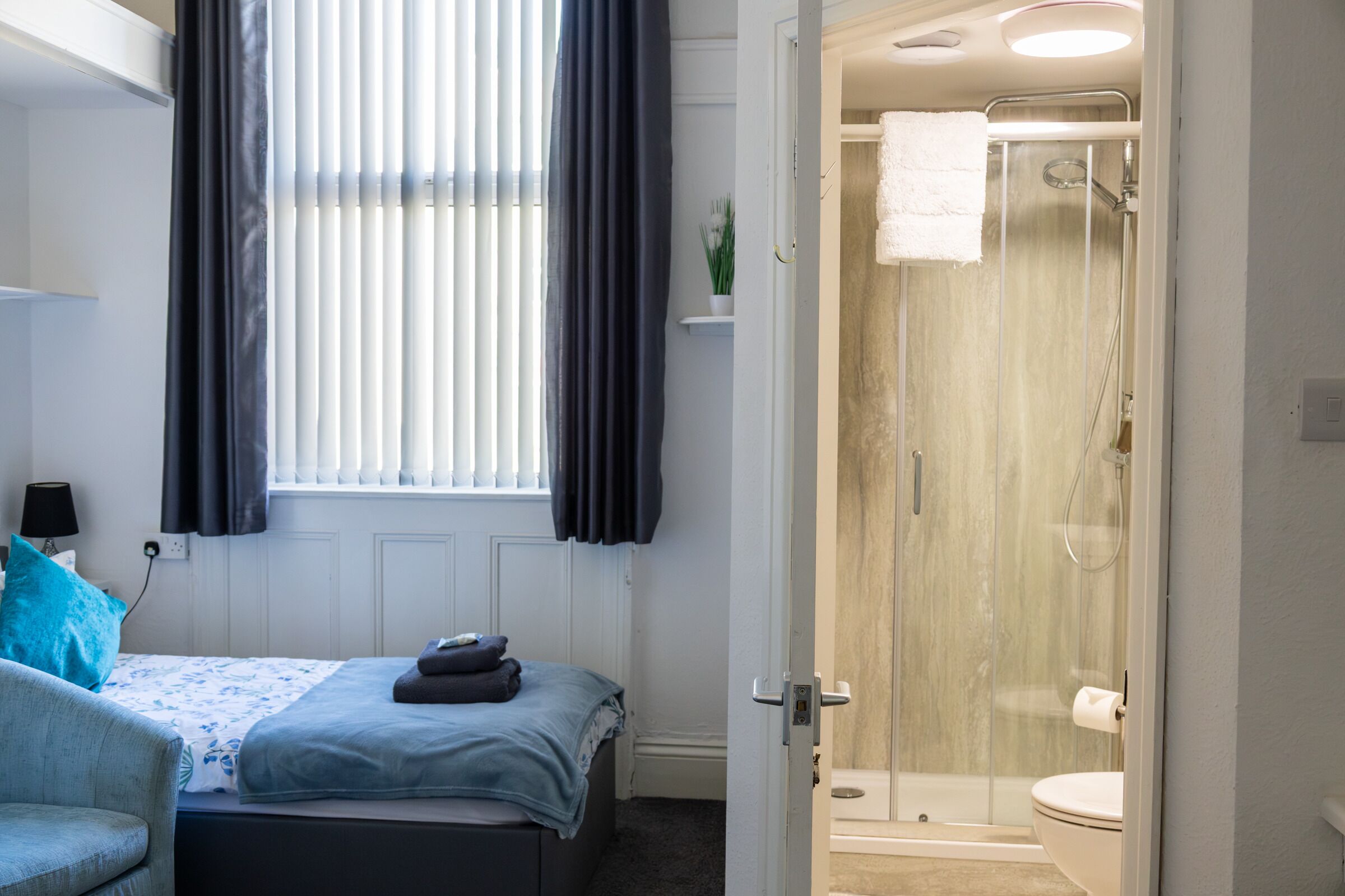 A bedroom with a bed, a chair, a window with curtains, and a bathroom with a shower and toilet.