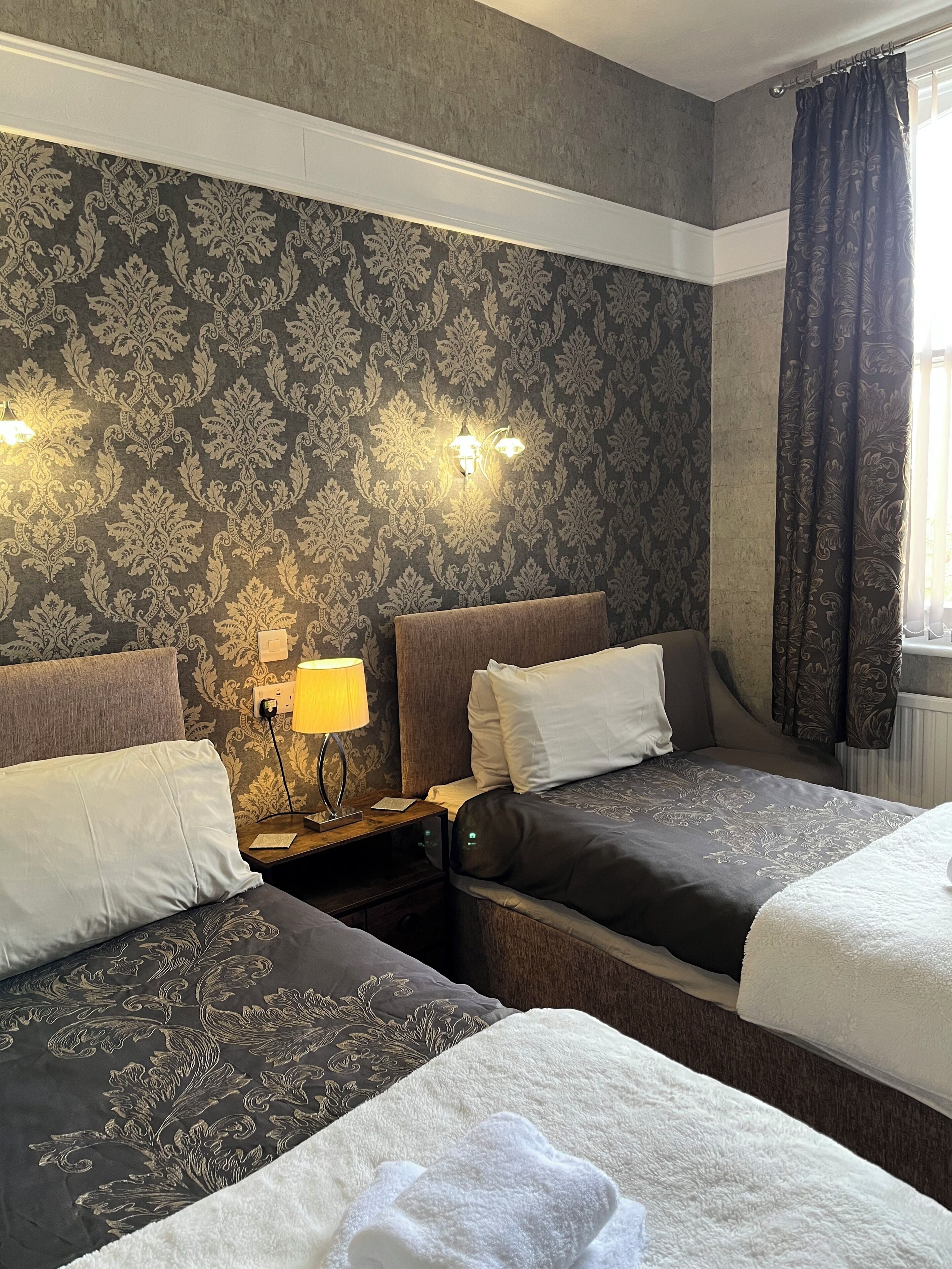 A hotel room with two beds, a nightstand with a lamp, patterned wallpaper, and a window with curtains.