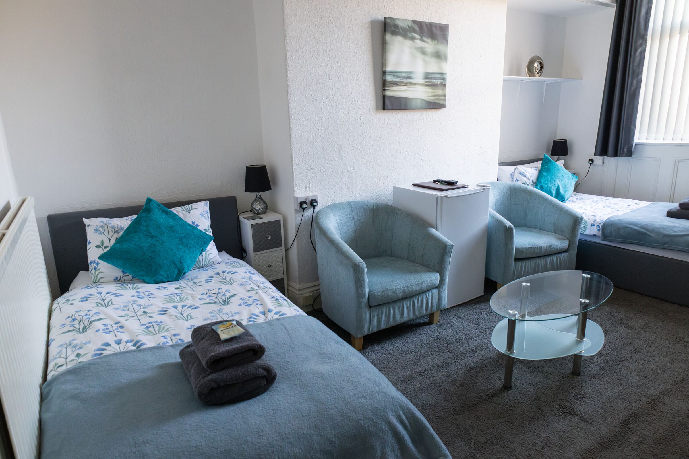 A hotel room with two beds, a chair, a small table, and a painting on the wall.
