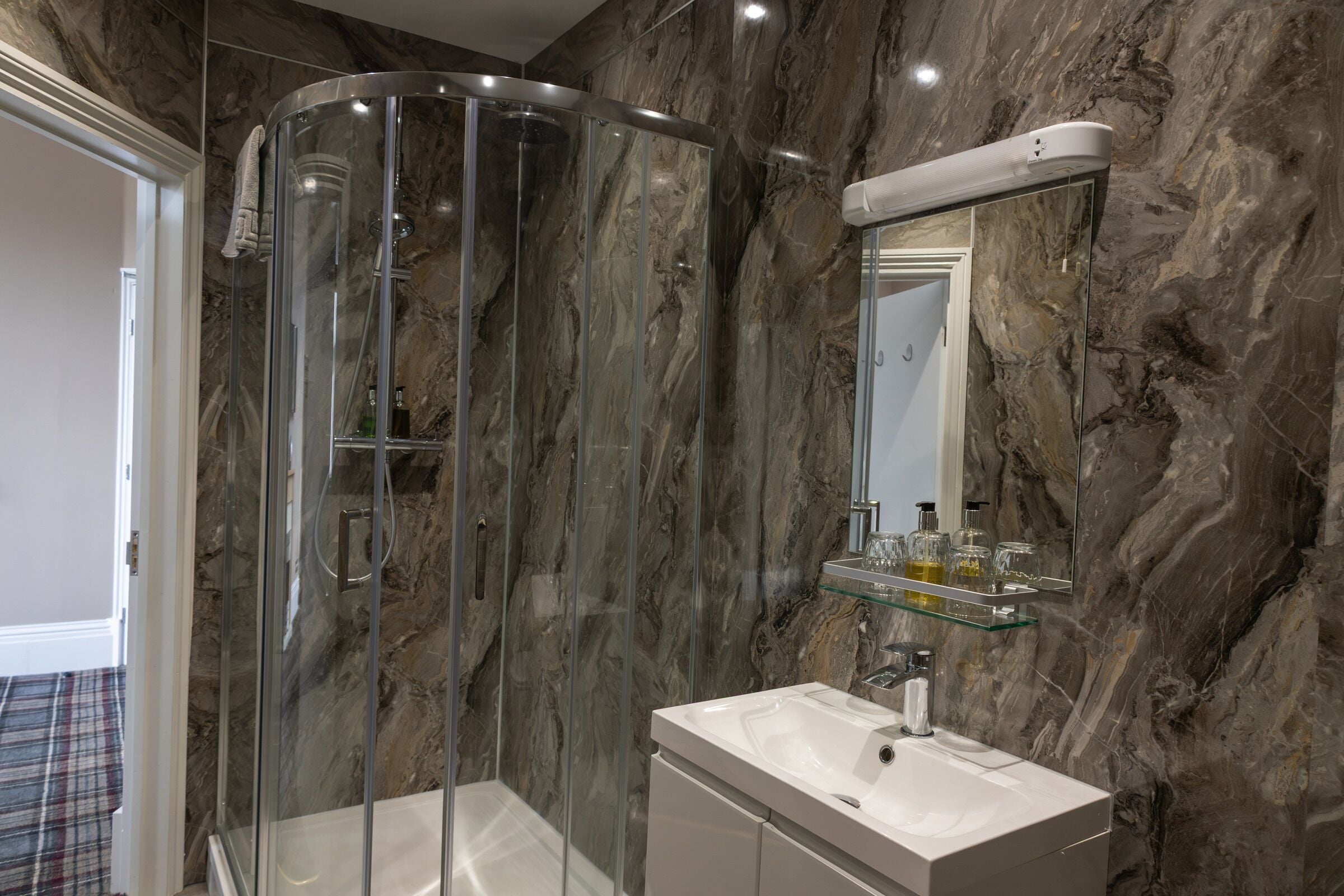 Un baño moderno con ducha de vidrio, pared de mármol, lavamanos blanco y repisa de vidrio con artículos de aseo.