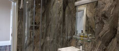 Un baño moderno con ducha de vidrio, pared de mármol, lavamanos blanco y repisa de vidrio con artículos de aseo.